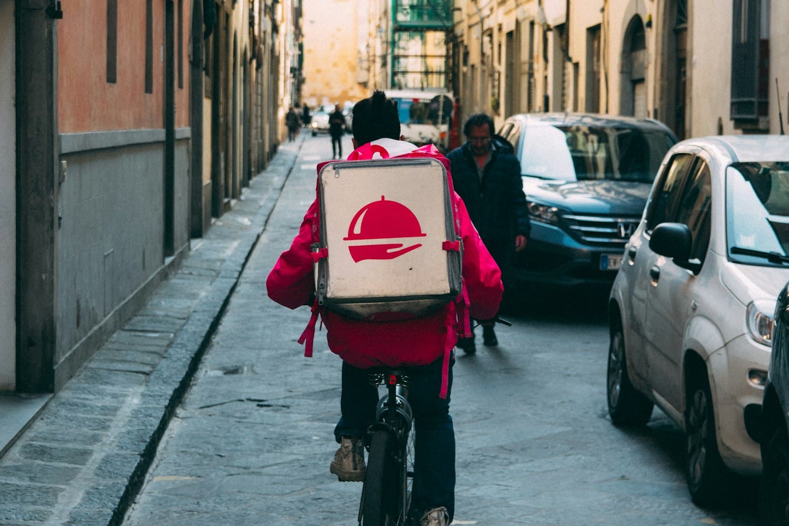A food delivery rider cycling down a narrow street with a food box on their back